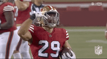 Sports gif. Talanoa Hufanga from the San Francisco 49ers runs across the field in slow motion looking up and signing "I love you" to someone above. 