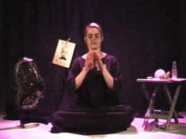 Woman sits crossed legged on the floor, surrounded by a large geode and occult crystals of all shapes and sizes. She makes a rainbow with her hands, sparkles trailing the motion in an arc, and dramatically drags out the second syllable of "Magic," which appears as text.
