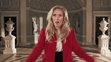 Reality TV gif. A blonde woman from the British TV show, Horrible Histories, in a bright red blazer raises her hands near her head and looks directly at us as she says excitedly, "Awesome!" which appears as text