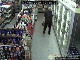 Video gif. Surveillance footage inside a convenience store shows a man carrying a six-pack walking and then falling over sloppily onto his back, apparently drunk.
