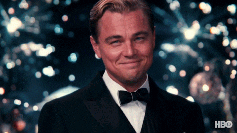 Gif s Leonardem DiCapriem v roli Gatsbyho, zvedajícím sklenici šampaňského k přípitku na pozadí vybuchujících ohňostrojů.