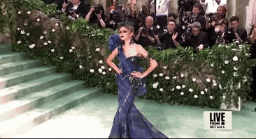 Met Gala 2024 gif. Zendaya poses with both hands on her waist as she sweeps her head down for a dark, ominous stare. She's wearing a midnight blue and dark teal dress with a full tulle puffed sleeve over her right shoulder and net mesh and tulle feathered fascinator. Her eye makeup makes a statement with heavy black eyeliner and bright red eyeshadow.