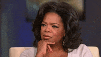 Celebrity gif. Oprah sits with her hand under her chin, frowning slightly and blinking.