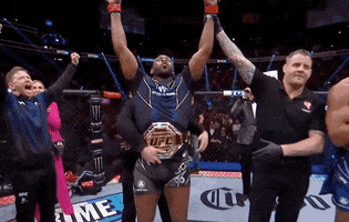 Video gif. Jon Jones holds up his hands in victory as he stands in the middle of the UFC ring as a UFC belt is tied around his waist. He yells, "Wooo!'