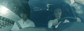 Video gif. Mother drives a car with her daughter in the front seat as we see the reflection of tall city buildings in the windshield.