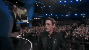 Oscars 2024 gif. In disbelief, Robert Downey Jr receives his Oscars trophy for Best Supporting Actor from Ke Huy Quan. He grabs the trophy with one hand and shakes hands firmly with Tim Robbins. 