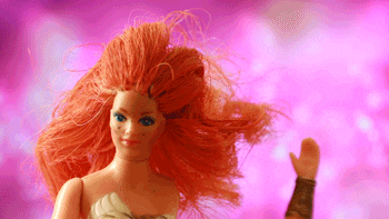 Video gif. Against a sparkly pink background, an 80s red headed female action figure stares at us, and its hand "waves" at us from bottom of frame.