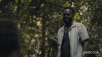 TV gif. William Jackson Harper as Noah on The Resort. They're in a jungle and he's smiling at them before promptly passing out, falling on his back.