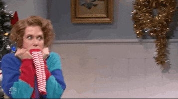 SNL gif. Kristen Wiig as Sue, wriggles next to a Christmas tree, hands clutching her turtleneck, stocking over her mouth, trying but failing to contain her exuberant excitement.