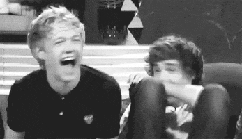 Celebrity gif. Louis Tomlinson from One Direction pulls his knees to his chest while Niall Horan falls back laughing on a couch.