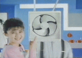 TV gif. As a woman points at an electric fan suspended from the ceiling, we zoom in to the fan's right. A man in suspenders leans out from behind the fan and mischievously covers his mouth.