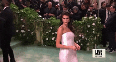 Met Gala 2024 gif. We zoom in on Kaia Gerber wearing a white strapless floor-length Prada gown featuring a paillette pattern of large white sequins with a straight neckline. Her hair is styled in a side-part bouffant. This is paired with a delicate diamond pendant necklace.