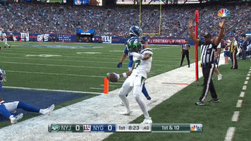 Sports gif. Garrett Wilson of the New York Jets does a celebratory dance on a football field, leaning back to wind up then throw a football into the air as his teammates rush over to him and a referee holds up the sign for touchdown. 