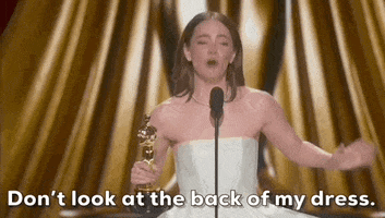 Oscars 2024 GIF. While crying, Emma Stone flails her arms and says, "Don't look at the back of my dress" while slightly turning and pointing to the back of her dress. 