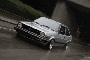 Video gif. Silver Volkswagen Jetta A2 driving fast down a highway on an overcast day.