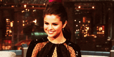Celebrity gif. Selena Gomez smiles, facing downward as if flattered, at a talk show appearance.