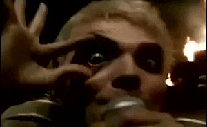 Music video gif. Gerard Way of My Chemical Romance holds his eye open wide as fire shoots off in the background. 