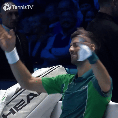 Video gif. Novak Djokovic sits on the sidelines of a match next to his tennis bag. He has a serene smile on his face as he conducts the crowd with his hands.