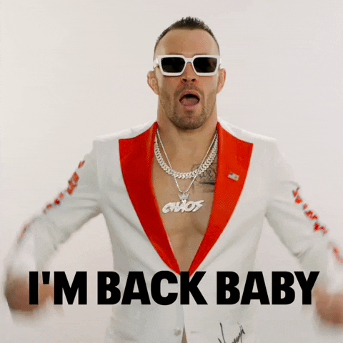 Video gif. UFC fighter Colby Covington wears a white suit, white sunglasses, and a silver chain necklace over his bare chest as he holds up his fists to make biceps, smiling with confidence as he says , "I'm back baby."