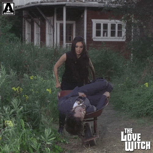 Movie gif. Samantha Robinson as Elaine in the Love Witch maneuvers through a yard driving wheelbarrow that holds Jeffrey Vincent Parise as Wayne, who looks dead.