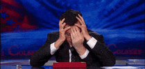 TV gif. Stephen Colbert as host of the Late Show rests his head in the palms of his hand. Two additional hands reach from below the desk to help hold his face.