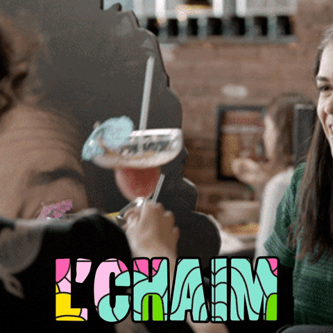TV gif. Abbi Jacobson and Ilana Glazer as Abbi and Ilana in Broad City, repeatedly raising, clinking, and sipping margarita glasses with little umbrellas. Colorful text, "l'chaim."