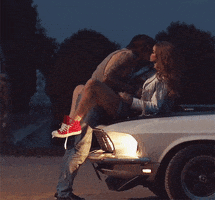 Music video gif. From the video for "Born to Die," Lana Del Rey sitting on the hood of a car as a man leans in, kissing her.