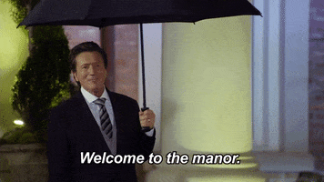 Reality TV gif. Martin Andrew as The Butler on Joe Millionaire: For Richer or Poorer. He greets contestants as they walk in, holding an umbrella open and saying, "Welcome to the manor."