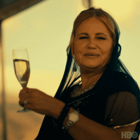 TV gif. Jennifer Coolidge as Tanya in The White Lotus smiles and raises a glass of wine. Text, "wee-hee!"