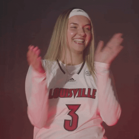 Video gif. Blonde woman wearing a Louisville Cardinals jersey leans forward, smiling and clapping energetically at us against a gray background strobing with red light. 