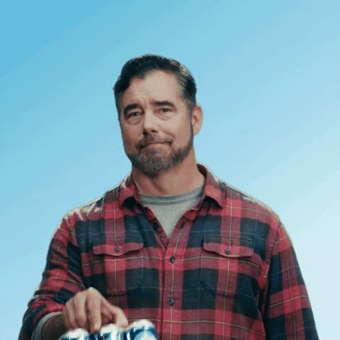 Sponsored gif. Gerald Downey in a red and black plaid shirt lifts a six pack of Busch Light beer out towards us, dipping his chin to give us a friendly, inviting expression like he's miming the question that appears on screen, "You in?"