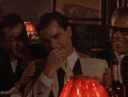 Movie gif. Ray Liotta as Henry Hill in Goodfellas sits at a table with a group of men. He opens his eyes and mouth wide in shock, holding his chest. He starts giggling and then bursts out cackling, laughing with the rest of the men.