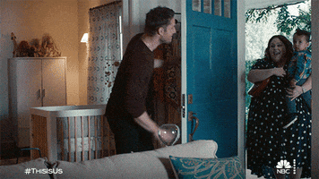 TV gif. Justin Hartley, as Kevin from This Is Us, opens the front door and greets Chrissy Metz as Kate, who walks in holding a toddler, followed by Chris Sullivan as Toby, who is carrying a baby car seat and gifts.