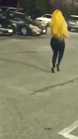Video gif. Shauna Brooks wearing a tight black catsuit with checkered racing stripes down the sides totters quickly across a parking lot in high heels, her long yellow hair flowing behind her as she struggles to pick up speed. 
