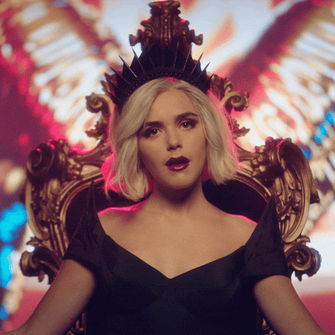 TV gif. Kiernan Shipka as Sabrina Spellman in the Chilling Adventures of Sabrina, sitting on a throne and winking.