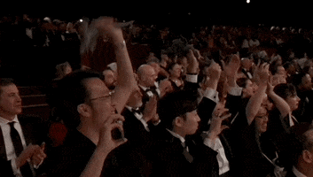 Oscars 2024 gif. The Godzilla Minus One team wins Visual Effects. Crew and cast members in the crowd stand and film the moment with their phones while clapping with their arms in the air. 