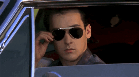 Movie gif. John Cusack as Lane in Better Off Dead sitting in a car with the window rolled down, pulls down his aviator sunglasses and winks.