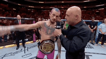 Sports gif. Sugar Sean O'malley is being interviewed by Joe Rogan at UFC 299 after his win and he says, “I love you, Joe Rogan.” Rogan responds, "I love you too," before patting his back and ending the interview.
