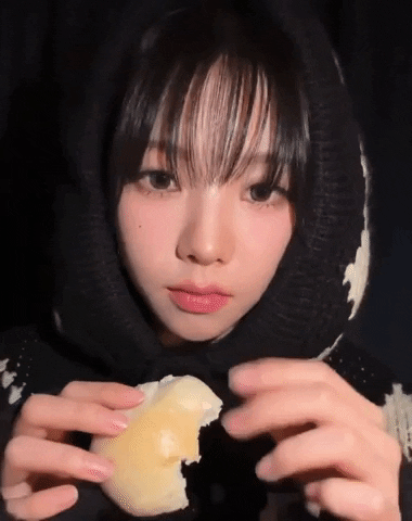 Celebrity gif. Closeup of Karina from the K-pop group aespa wears a black sweater hoodie tight around her face as she holds a half-eaten bun in one hand and makes a fist with the other, looking pretty serious like "I will punch you." 