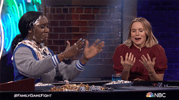 Reality TV gif. Kristen Bell on Family Game Fight cringes and covers her face to laugh as a woman next to her laughs with pie cream covering her head and chest.
