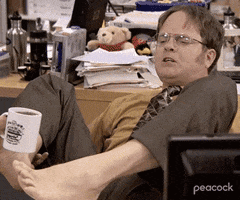 The Office gif. Rainn Wilson as Dwight sits at his desk holding a cup of coffee with his bear feet. He tries to pull it closer to himself to take a drink but it tilts as it comes closer and spills out all over his crotch. He freaks out, sticking out his tongue as it does.