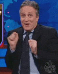 TV gif. Jon Stewart on the Daily show sits at his desk squirming around excitedly. He claps like he’s a baby and stares at us with a fake excited look on his face. 