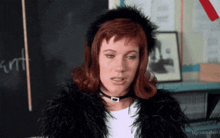 Movie gif. Elisa Donovan as Amber Mariens in Clueless standing in front of a chalkboard and wearing a feathered headband and coat, makes a "W" sign with her fingers and spits out the word, "Whatever." She's over it.