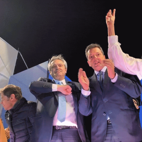 Video gif. Two Argentinian politicians dance on stage. One man gives two thumbs up while the other man bumps his chest with his fist and points at us.