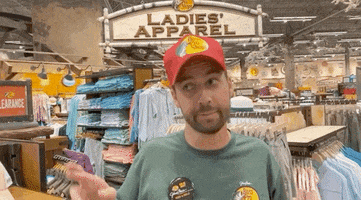 Celebrity gif. John Crist, wearing a Bass Pro Shop hat in a Bass Pro Shop, crosses his fingers and knocks on wood as he says, "Hopefully."