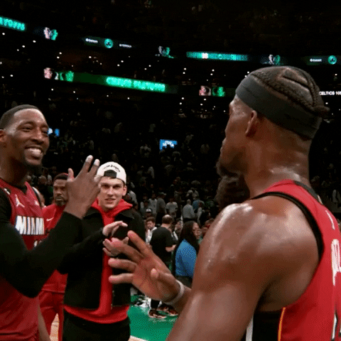 Sports gif. Slow motion clip of Jimmy Butler and Bam Adebayo from the Miami Heat walking towards each other on court as they slap backhands and then touch foreheads, looking confident and proud. 