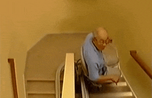 Video gif. An old man rides a chair lift assist down the stairs.