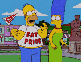 The Simpsons gif. Marge stands next to Homer who's wearing a "Fat Pride" shirt and holding a flag that reads, "Girth." He places a hand on his belly as his stomach slowly grows and protrudes from the bottom of his shirt. He lowers the flag. 