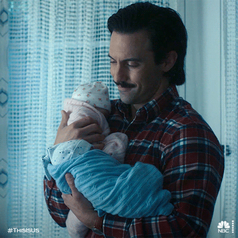 TV gif. Milo Ventimiglia as Jack from This Is Us proudly holds two small babies: one in a pink blanket and one in a blue blanket.