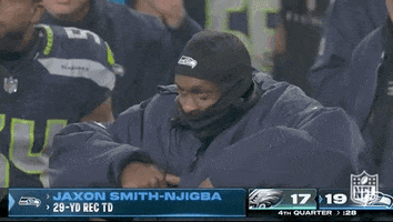 Sports gif. Jon Smith-Njigba, an American football player, is watching the game from the sidelines, wearing a helmet liner that's pulled up over his mouth so you can only see his eyes. He bobs his head up and down and looks pleased as he holds his elbows out like eagle wings. 